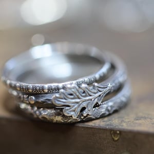 Puede incluir: Tres anillos de plata apilados. El anillo superior presenta un diseño detallado de hoja de roble. Los anillos están sobre una superficie marrón.