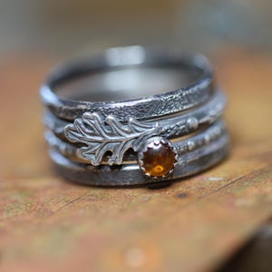 May include: A set of four silver rings featuring an oak leaf design and a small amber gemstone. The rings are stacked on top of each other and have a textured finish.