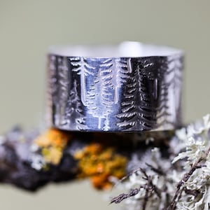 May include: A silver ring with a forest design. The ring has an etched pattern of trees around its band. The ring is displayed on a branch with lichen and moss, creating a natural aesthetic. The ring's width is approximately 1 cm.