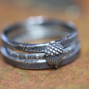 以下が含まれることがあります： 3 つのバンドとドングリのデザインが施されたシルバーリング。バンドはテクスチャ加工が施され、ドングリは細部まで作り込まれています。