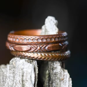 May include: A stack of three copper-toned metal rings, each featuring a distinct embossed pattern. The top ring has a scalloped edge, the middle ring displays a geometric diamond motif, and the bottom ring shows a braided or twisted rope texture.