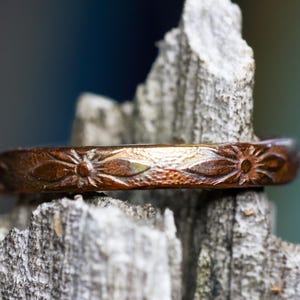 May include: A copper-toned metal ring featuring a repeating embossed floral pattern along its band. The surface of the ring displays a textured, metallic finish with dark brown accents within the recessed areas of the stamped flower designs.