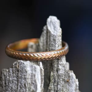 May include: A metallic ring with a warm, copper-toned finish and a repeating, slanted etched pattern encircling the entire band. The ring is positioned horizontally, resting against a jagged, light-colored piece of weathered, textured wood.