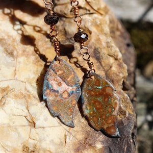 May include: Handmade copper leaf earrings with a unique patina. The earrings feature a leaf shape with a mix of brown, blue, and copper tones. Each earring has a dark brown bead and is suspended from a copper wire.