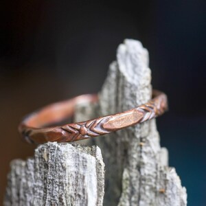 May include: A copper ring featuring a repeating etched pattern of small diamonds and lines along the outer band. The metal has a warm, reddish-brown metallic finish with visible texture from the engraving process, creating a distinct geometric design.