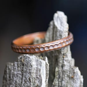 May include: A copper-toned metal ring featuring a repeating pattern of small, circular indentations along the outer band. The ring has a smooth, rounded interior surface and a textured, darkened finish within the circular impressions.
