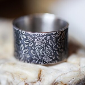 May include: A wide, silver-toned ring with an engraved design of an owl surrounded by leaves. The ring is set on a light-colored, textured surface. The ring's band is approximately 1 inch wide.