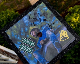 May include: A black graduation cap with a custom design featuring a rapper sitting on a building with the words "There's beauty in the struggle" and a crown. The cap also has a green street sign that says "Class of 2025".