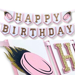 May include: A two-tier birthday banner featuring white pennants with pink borders. The letters spell HAPPY BIRTHDAY in gold glitter. Each end of the top row features a pink hockey puck graphic with black and gold glitter flames on a white background.