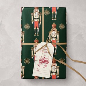 Puede incluir: Regalo envuelto con papel verde oscuro con un patrón repetido de soldados cascanueces y copos de nieve dorados. Una etiqueta de regalo con "Merry Christmas" y una ilustración de Papá Noel está atada con una cinta dorada.