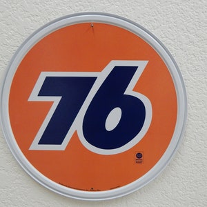 May include: Round orange and white metal sign with the number 76 in navy blue. The sign has a white border and a small hole at the top for hanging. The sign is a vintage style.