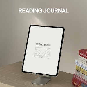 May include: A white digital tablet with a black screen displaying the text "READING JOURNAL" and a stylized image of an open book. The tablet is on a stand and there are several books stacked behind it.
