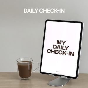 May include: A white tablet with the text "MY DAILY CHECK-IN" displayed on the screen. The tablet is on a silver stand. A glass mug with brown liquid is on a wooden surface.