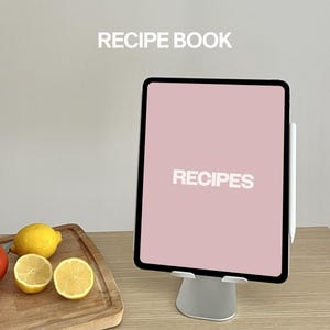 May include: A white tablet stand with a pink screen displaying the word "RECIPES". The tablet is on a wooden cutting board with lemons and tomatoes.
