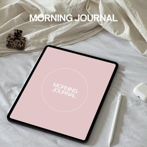 Puede incluir: Una tableta rosa con un círculo blanco que dice "MORNING JOURNAL" sobre una colcha blanca. Un lápiz blanco y una funda blanca para auriculares inalámbricos también están sobre la colcha.