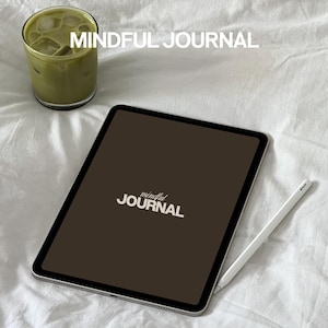 Op de afbeelding: Een bruine digitale tablet met een witte stylus en de tekst "mindful JOURNAL" op het scherm. De tablet ligt op een wit beddengoed met een glas groene ijsthee op de achtergrond.