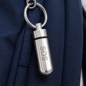 Könnte beinhalten: Ein zylindrischer Kapselanhänger aus silberfarbenem Metall mit der Gravur SOS auf der Vorderseite. Die Kapsel ist an einem runden Metallring befestigt, der am dunkelblauen Stoff-Reißverschlussanhänger eines strukturierten marineblauen Rucksacks hängt.