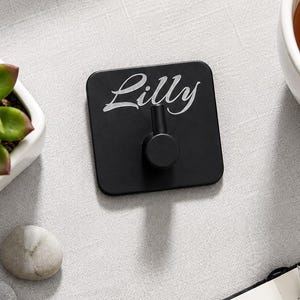 Könnte beinhalten: Ein schwarzer quadratischer Wandhaken mit dem Namen "Lilly" in weißer Schrift. Der Haken befindet sich auf einer hellgrauen Oberfläche, neben einer kleinen Sukkulente in einem weißen Topf, zwei glatten Steinen, einer Tasse Tee und einem Notizbuch mit einem Stift.