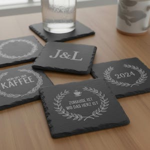 Puede incluir: Un conjunto de posavasos cuadrados de pizarra gris oscuro con varios diseños y textos grabados en blanco. Un posavasos muestra las iniciales "J&L", otros muestran frases y el año "2024". Un vaso de agua y una taza son visibles en el fondo.