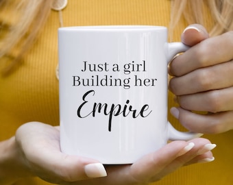 Pode incluir: Caneca de cerâmica branca com o texto "Just a girl Building her Empire" em escrita preta. A caneca é segurada por uma pessoa vestindo uma camisa amarela. A caneca tem uma alça no lado direito.