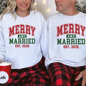 May include: Two ash grey crewneck sweatshirts featuring the red and green text MERRY AND MARRIED EST. 2025 printed on the front. The sweatshirts have long sleeves and a classic fit, paired with red and black buffalo plaid patterned bottoms.