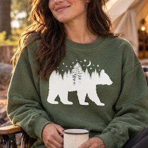 May include: An olive green crewneck sweatshirt featuring a white graphic print of a bear silhouette filled with a forest of pine trees, a crescent moon, and stars. The long-sleeved pullover has a ribbed neckline, cuffs, and hemline.