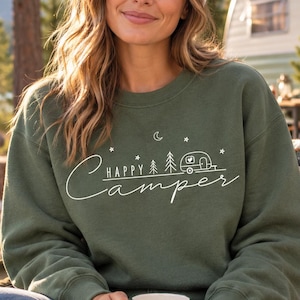 May include: An olive green crewneck sweatshirt featuring white screen-printed text that reads Happy Camper, accompanied by graphic illustrations of a small travel trailer, pine trees, a crescent moon, and stars. A white mug with a black tree print.