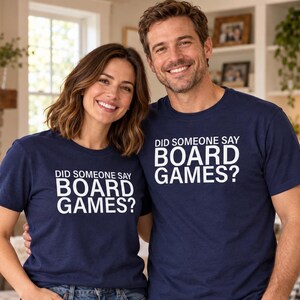 May include: Two navy blue short-sleeve t-shirts featuring the white text DID SOMEONE SAY BOARD GAMES? printed on the chest. The shirts are made of a solid-coloured fabric and have a crew neckline, paired with blue denim jeans.