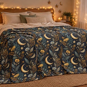 May include: A dark blue throw blanket featuring a repeating pattern of golden crescent moons, small stars, and botanical illustrations of yellow flowers and green leaves. The fabric has a soft texture and covers the surface of a bed.