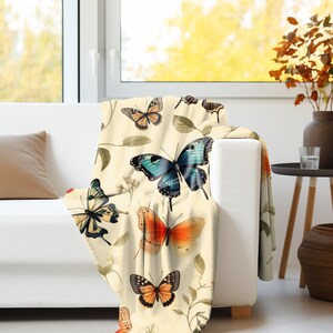 Op de afbeelding: Een cr&egrave;mekleurige plaid met een herhalend patroon van diverse vlinders in tinten blauw, oranje en zwart, afgewisseld met delicate botanische illustraties van bladeren en kleine steeltjes over het gehele oppervlak van de stof.