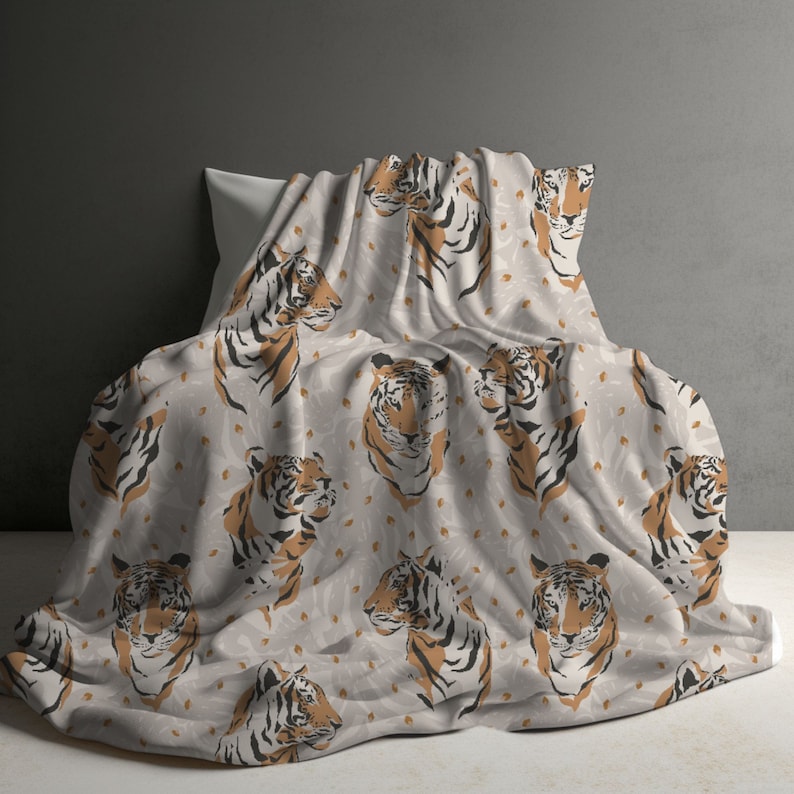 May include: A light beige blanket featuring a repeating pattern of tiger heads in shades of orange, black, and white. The tiger illustrations are arranged in a grid-like layout across the fabric, which is draped over a pillow to show the design.