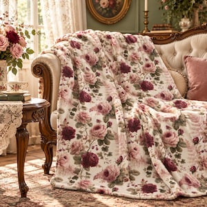 May include: A soft, plush throw blanket featuring a repeating pattern of deep burgundy and soft pink roses with green leaves on a cream-colored background, draped over the arm and seat of a tufted, curved wooden sofa.