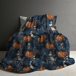 May include: A soft blanket featuring a dark navy blue background with a repeating pattern of orange pumpkins, dark blue foliage, and small white flowers. The fabric is draped over a pillow, displaying the detailed botanical and seasonal print.