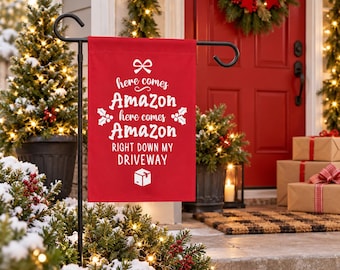 May include: A red rectangular garden flag hanging on a black metal stand. The flag features a white bow icon, white holly sprigs, and the text: here comes Amazon here comes Amazon RIGHT DOWN MY DRIVEWAY, with a white shipping box icon below.