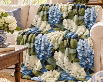 May include: A soft throw blanket featuring a dense, repeating pattern of blue and white hydrangea flower clusters interspersed with large, detailed green leaves against a dark background, draped over the arm of a beige sofa.