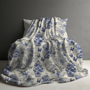 May include: A cream-colored blanket featuring a repeating pattern of blue ginger jars, floral arrangements, and butterflies. The fabric displays a detailed toile-style print with deep blue ink illustrations against a light, neutral background.