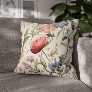 May include: A cream-coloured throw pillow with a floral pattern featuring pink, red, blue, and yellow flowers. The pillow has a white background and is made of a soft, comfortable fabric.