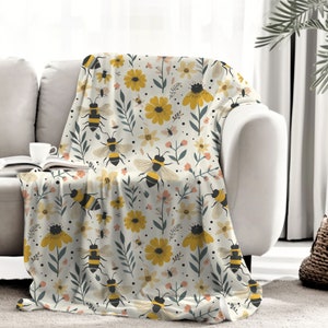 May include: A soft, white throw blanket with a repeating pattern of yellow flowers, green leaves, and black and yellow bees.