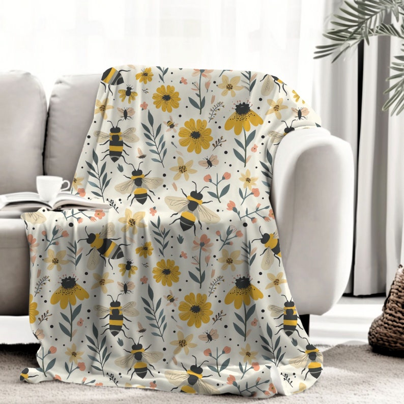 May include: A soft, white throw blanket with a repeating pattern of yellow flowers, green leaves, and black and yellow bees.