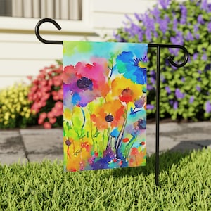 May include: A vibrant garden flag featuring a watercolor painting of colorful flowers in shades of pink, orange, yellow, and blue. The flag is displayed on a black metal stand, set against a backdrop of green grass and flowering bushes.