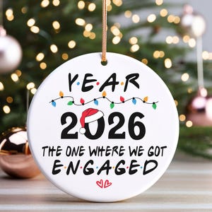 May include: Round white ceramic Christmas ornament with the text "YEAR 2026 THE ONE WHERE WE GOT ENGAGED". The number 2026 has a Santa hat. A string of colourful lights is above the numbers. The ornament hangs from a gold string.