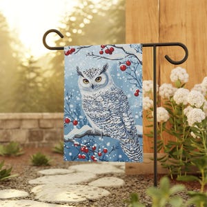 May include: A garden flag featuring a snowy owl perched on a snow-covered branch with red berries against a light blue background. The flag is displayed on a black metal stand, perfect for outdoor decor.