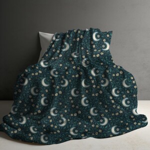 May include: A dark teal blanket featuring a repeating pattern of white crescent moons, small light-colored flowers, and dark green leafy vines. The fabric is draped over a pillow, displaying a consistent botanical and celestial print throughout.