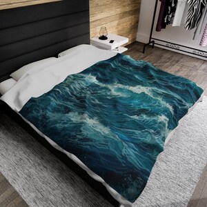 May include: A plush bed blanket featuring a detailed, high-contrast print of deep blue ocean waves with white foam crests. The design covers the top surface of the blanket, which is draped over a bed with white bedding and a black headboard.