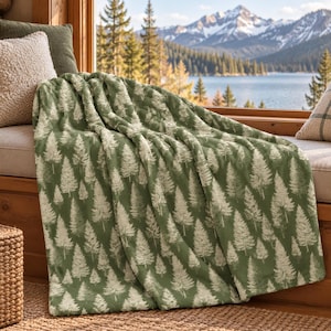 Op de afbeelding: Een zachte, pluche plaid in een diepe saliegroene kleur met een herhalend patroon van witte dennenbomen. De stof heeft een getextureerd, pluizig uiterlijk met zichtbare plooien en draperingen, die de gedetailleerde boomprint goed weergeven.