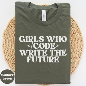 Puede incluir: Camiseta verde militar con el texto blanco "GIRLS WHO </CODE> WRITE THE FUTURE". La camiseta se muestra sobre una estera circular tejida. La marca "CANVAS" es visible en la etiqueta. La camiseta está hecha de un material suave.