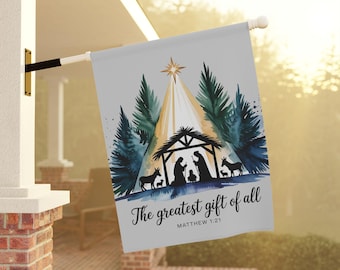 Può includere: Una bandiera grigia da esterno con una scena della Natività, una stella, alberi e il testo "The greatest gift of all" e "Matthew 1:21". La bandiera è appesa a un palo bianco con una staffa nera.