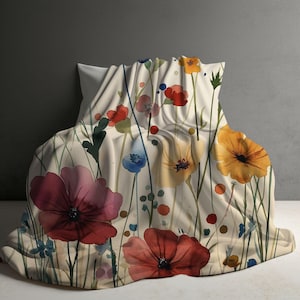 May include: A cream-colored blanket with a watercolor floral design. The blanket features large, colorful flowers in shades of red, pink, yellow, and blue, along with green stems and leaves. The blanket is draped over a white pillow.
