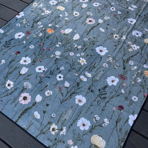 May include: A blue and white floral patterned picnic blanket with a white border. The blanket features a variety of white flowers and green stems on a blue background.