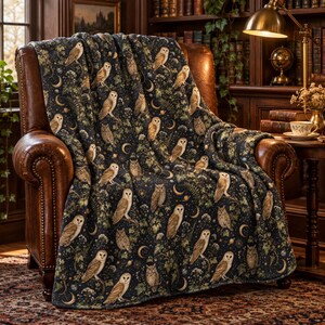 May include: A dark navy blue throw blanket featuring a repeating pattern of brown owls, crescent moons, stars, and green foliage. The blanket is draped over a brown leather armchair, showcasing a detailed print of various owl species.